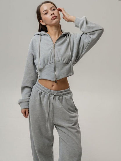Donna in tuta grigia, felpa con cappuccio e pantaloni larghi, posa su sfondo neutro. Abbigliamento casual, moda comoda, stile sportivo.