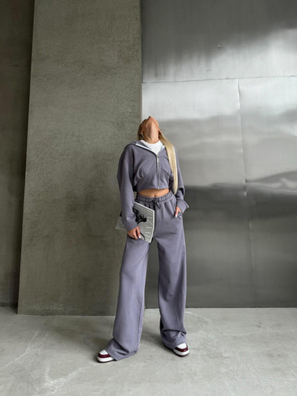Donna in tuta grigia oversize con cappuccio, pantaloni larghi e scarpe da ginnastica bianche e rosse, posa in un ambiente industriale minimalista.