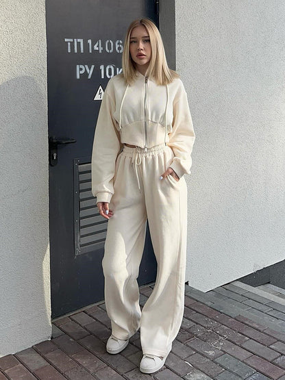 Giovane donna in tuta sportiva beige, con felpa corta e pantaloni larghi, posa davanti a una porta grigia. Look casual e moderno.
