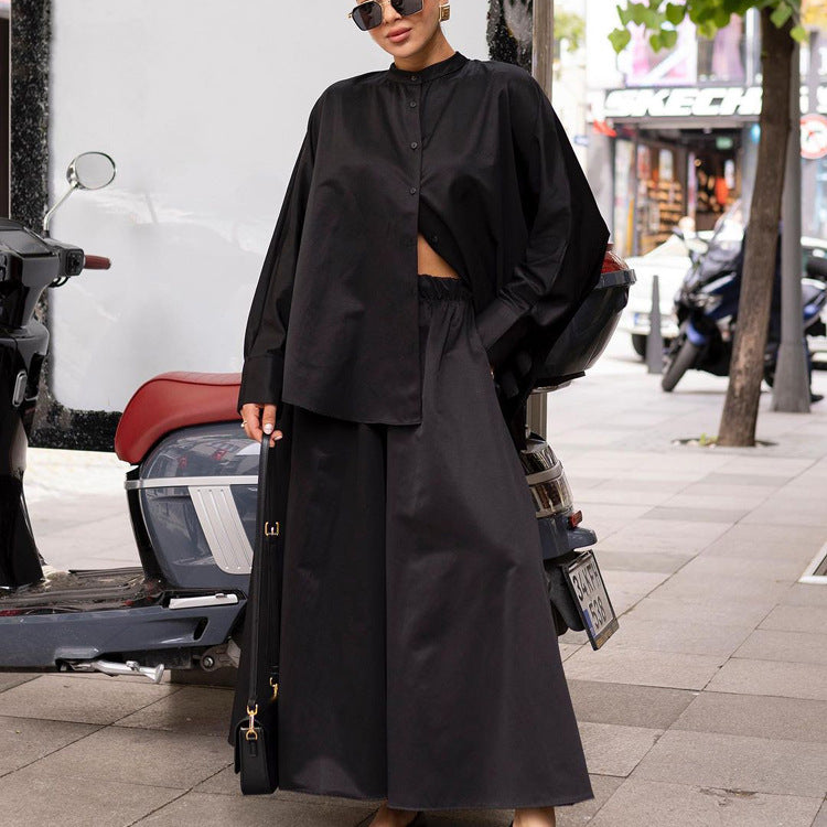 Donna con abito nero oversize, camicia e gonna lunga, posa accanto a scooter su marciapiede cittadino. Moda urbana, stile elegante.