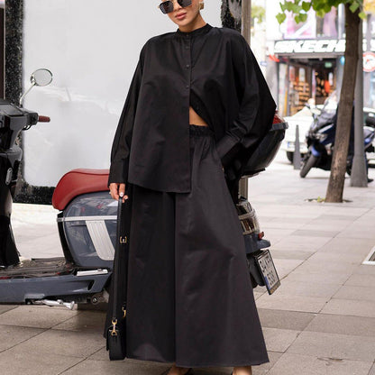 Donna con abito nero oversize, camicia e gonna lunga, posa accanto a scooter su marciapiede cittadino. Moda urbana, stile elegante.