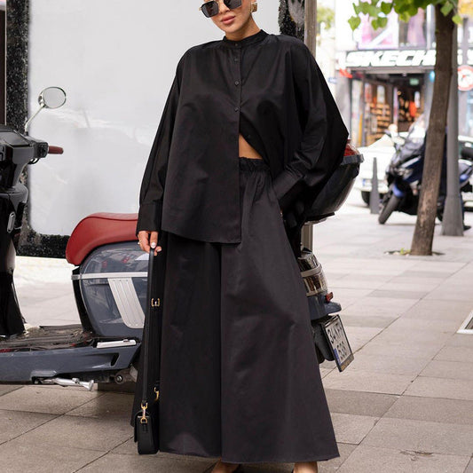 Donna con abito nero oversize, camicia e gonna lunga, posa accanto a scooter su marciapiede cittadino. Moda urbana, stile elegante.