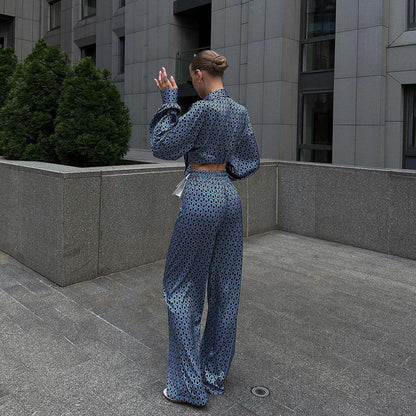 Donna di spalle in tuta elegante blu con motivo geometrico, cammina su marciapiede urbano. Edificio moderno grigio sullo sfondo. Moda urbana chic.