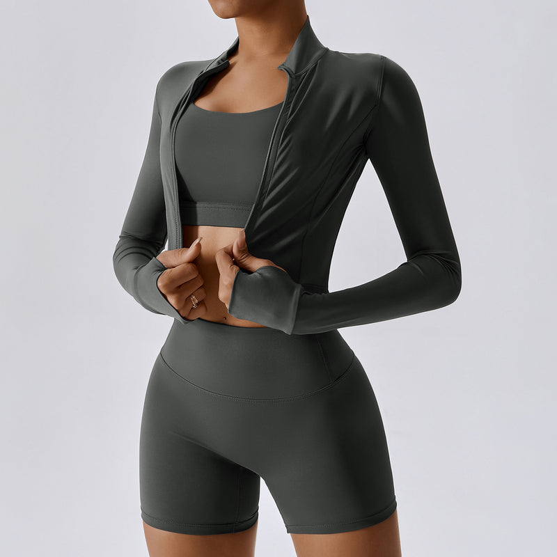 Donna in abbigliamento sportivo grigio scuro, con top e pantaloncini aderenti, apre una giacca leggera. Fitness, moda activewear, abbigliamento palestra.