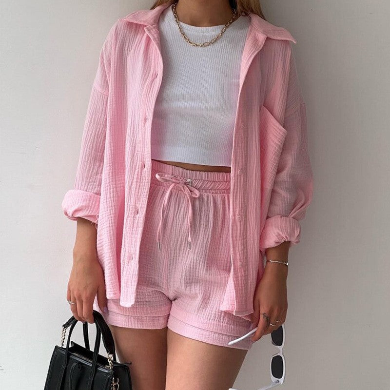 Donna con camicia rosa oversize, top bianco e pantaloncini coordinati, tiene una borsa nera e occhiali da sole. Moda estiva casual elegante.