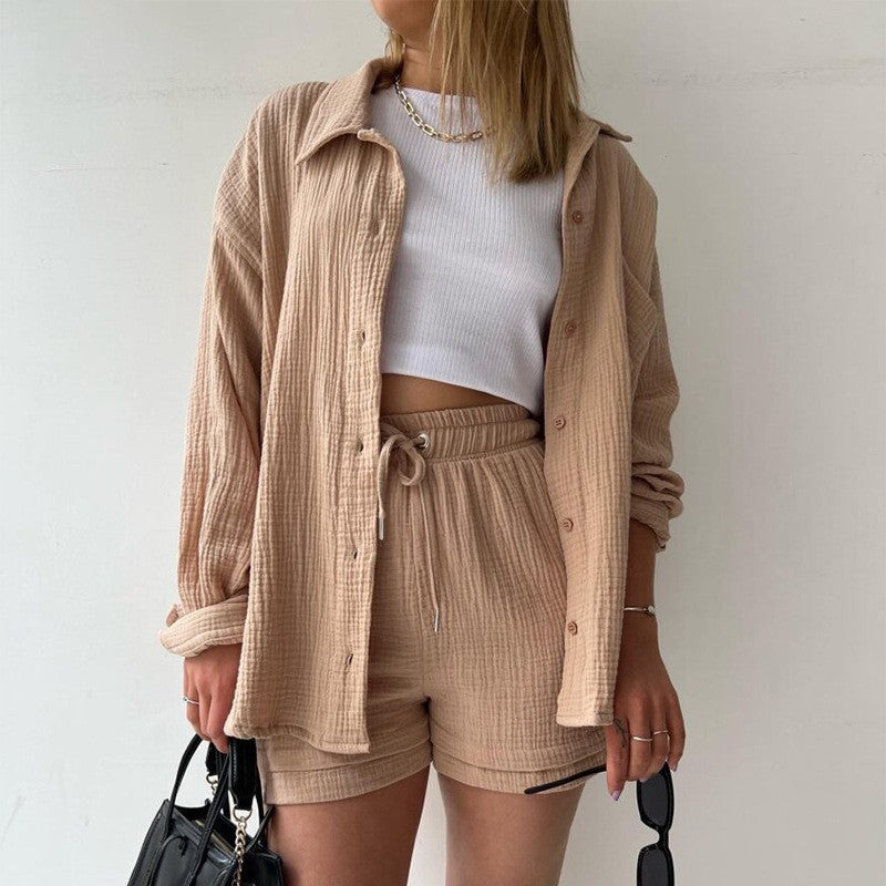 Donna con camicia beige e pantaloncini coordinati, top bianco, collana dorata, borsa nera. Moda casual estiva, abbigliamento femminile trendy.