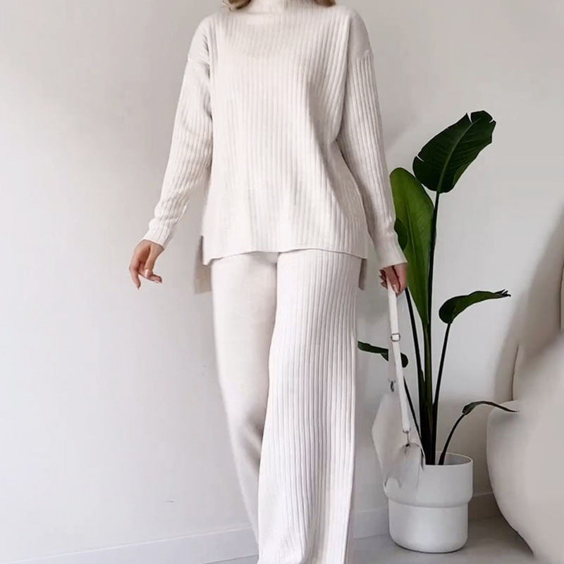Donna in abbigliamento loungewear bianco, maglione e pantaloni a coste, con borsa bianca. Pianta verde sullo sfondo. Moda casual elegante.