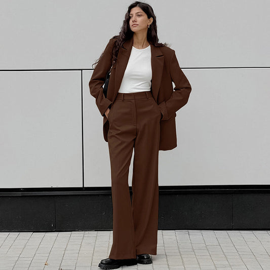 Donna in abito marrone elegante, giacca e pantaloni, su sfondo grigio. Moda autunnale, stile business casual, tendenze abbigliamento femminile.