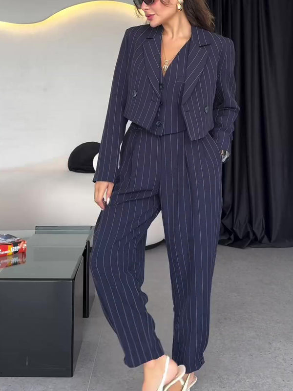 Donna in elegante completo gessato blu scuro, giacca corta e pantaloni larghi, indossa occhiali da sole e sandali bianchi, in un salotto moderno.