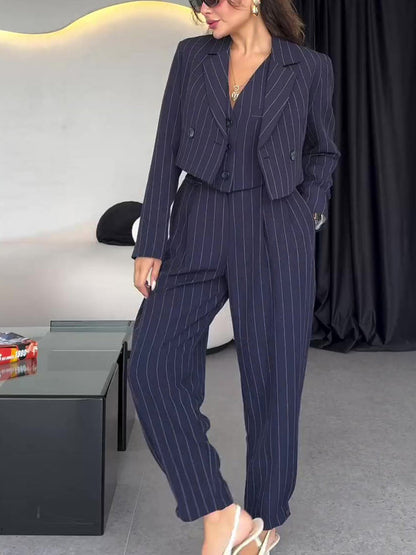 Donna in elegante completo gessato blu scuro, giacca corta e pantaloni larghi, indossa occhiali da sole e sandali bianchi, in un salotto moderno.