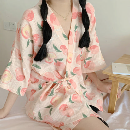 Kimono estivo donna con stampa pesche, tessuto leggero, maniche ampie, cintura in vita, seduta su letto, tavolino legno accanto.