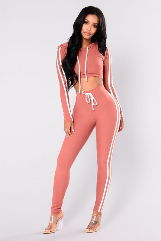 Donna in tuta sportiva rosa con strisce bianche, top corto con cappuccio e pantaloni a vita alta, indossa sandali trasparenti. Moda casual femminile.