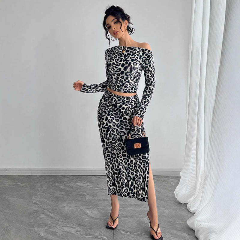 Donna in abito leopardato a spalle scoperte, con borsa nera e sandali, posa in una stanza moderna con tende bianche. Moda femminile elegante.