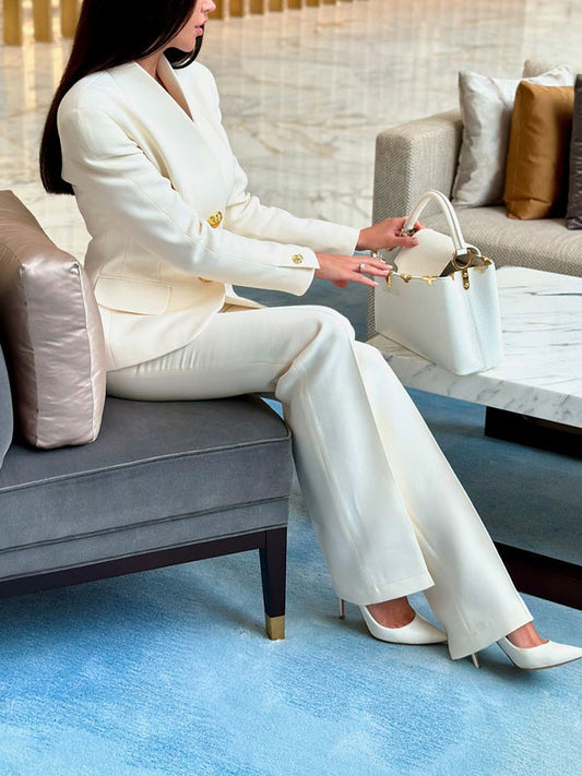Donna in elegante completo bianco seduta su divano moderno, con borsa bianca e scarpe abbinate, in un ambiente lussuoso e raffinato.