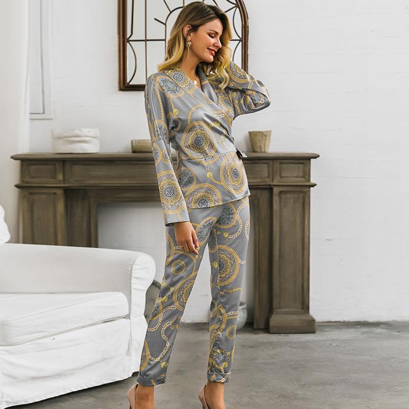 Donna in pigiama di seta grigio con motivi dorati, in piedi vicino a un camino in un salotto moderno. Moda notte elegante e confortevole.