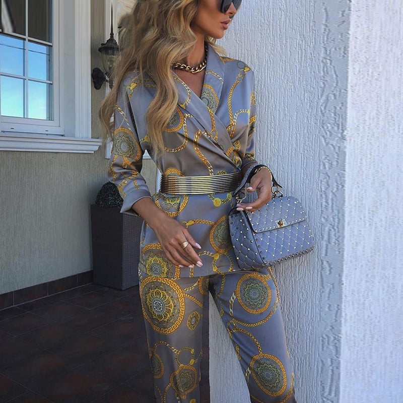 Donna elegante in tuta grigia con motivi dorati, borsa trapuntata grigia, collana dorata, occhiali da sole, posa su balcone moderno.