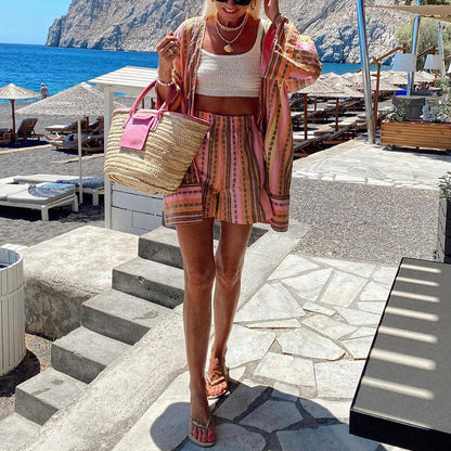Donna in spiaggia con abbigliamento estivo colorato, borsa di paglia e sandali. Mare e lettini sullo sfondo. Look estivo alla moda.