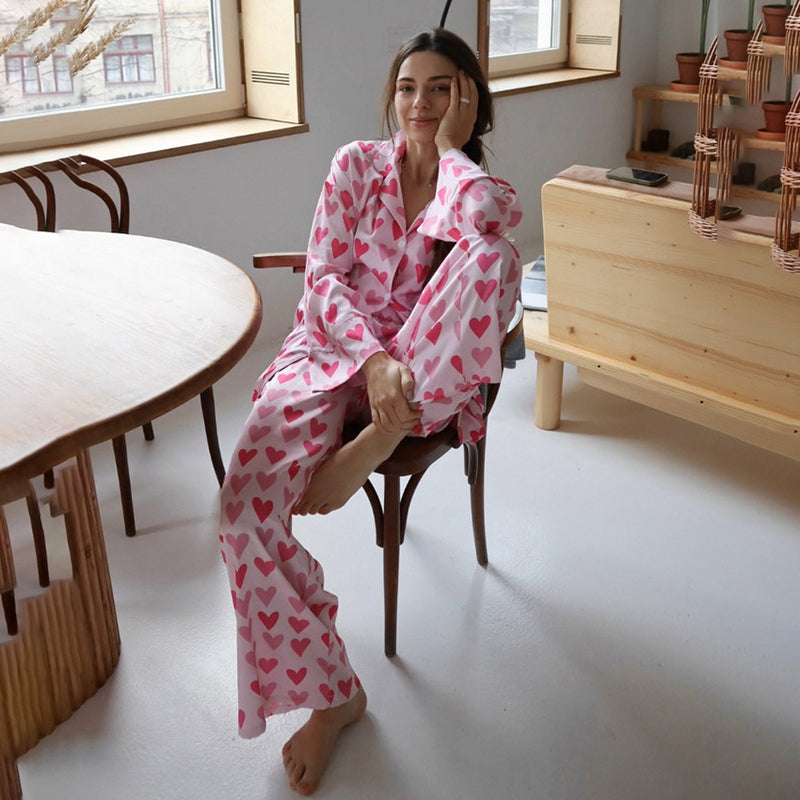 Donna seduta in pigiama rosa con cuori, in una cucina moderna. Tavolo in legno e finestre luminose sullo sfondo. Comfort e stile casual.