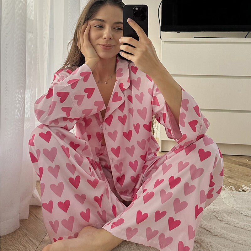 Donna in pigiama rosa con cuori rossi, seduta sul pavimento, scatta un selfie con smartphone. Moda notte, abbigliamento comodo, stile casual.