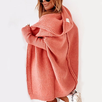 Donna con cardigan oversize rosa, cappuccio ampio, occhiali da sole, scarpe bianche. Moda autunnale, abbigliamento casual elegante.