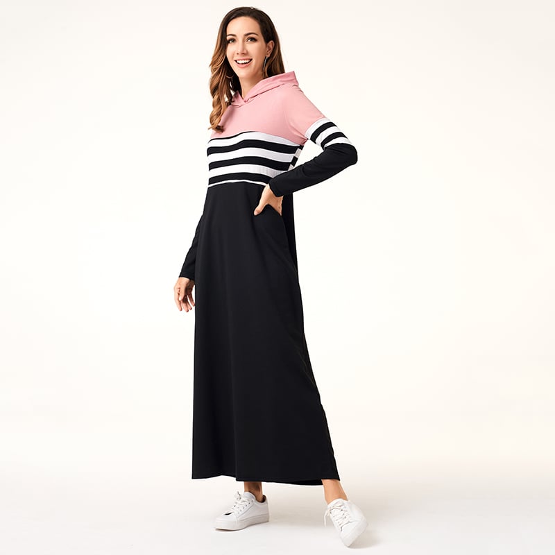 Donna in abito lungo a righe nere e bianche con cappuccio rosa, sorridente, in posa su sfondo bianco. Moda casual, abbigliamento femminile elegante.