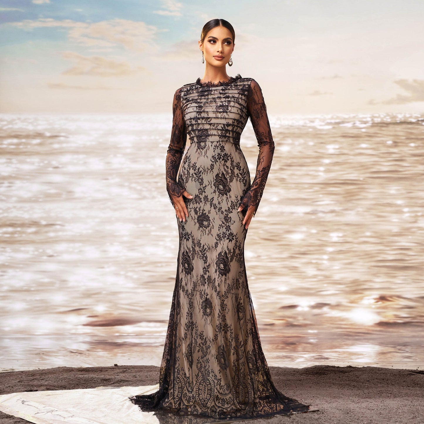 Donna in abito da sera lungo in pizzo nero, elegante e sofisticato, su sfondo di mare al tramonto. Moda femminile, abbigliamento di lusso, stile glamour.