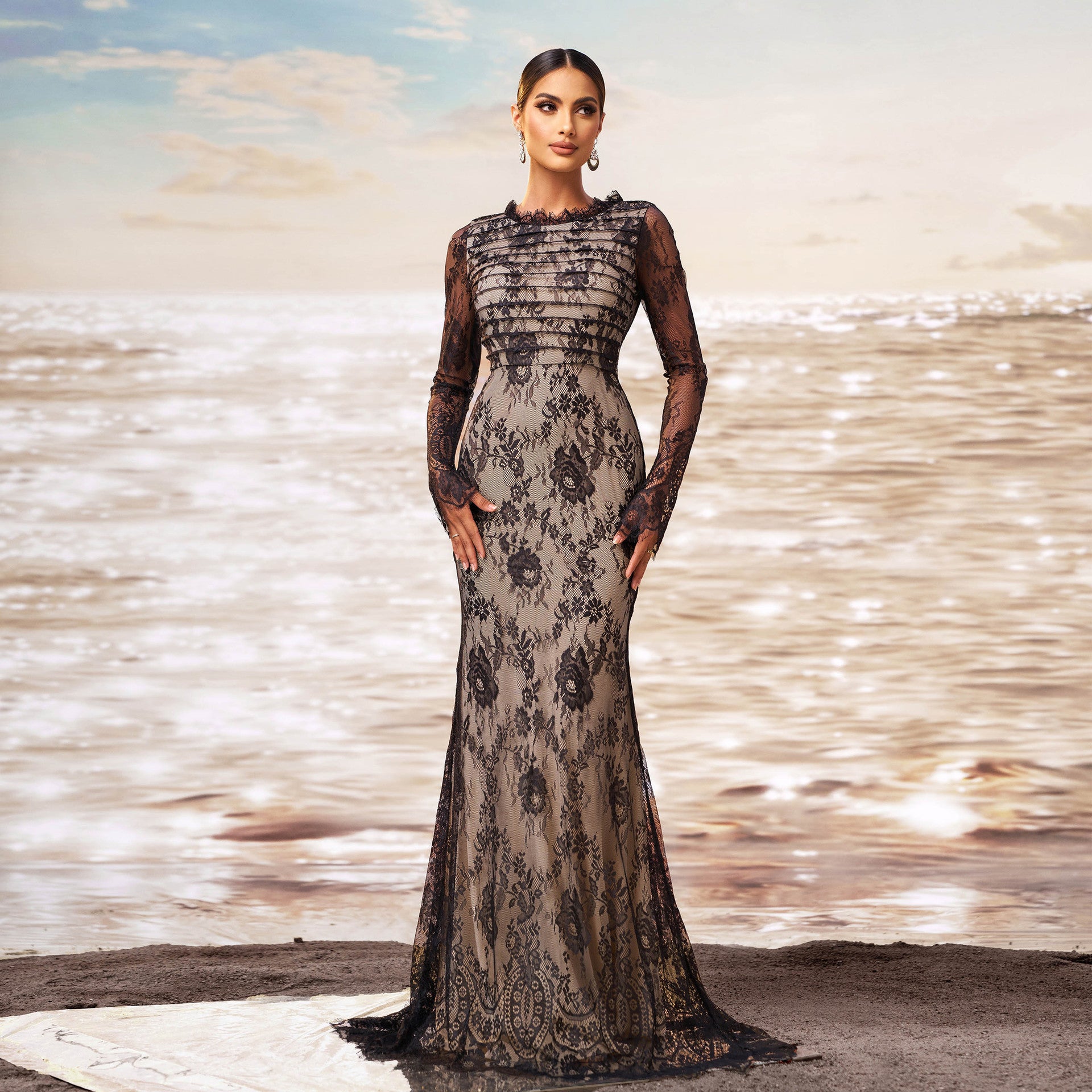Donna in abito da sera lungo in pizzo nero, elegante e sofisticato, su sfondo di mare al tramonto. Moda femminile, abbigliamento di lusso, stile glamour.
