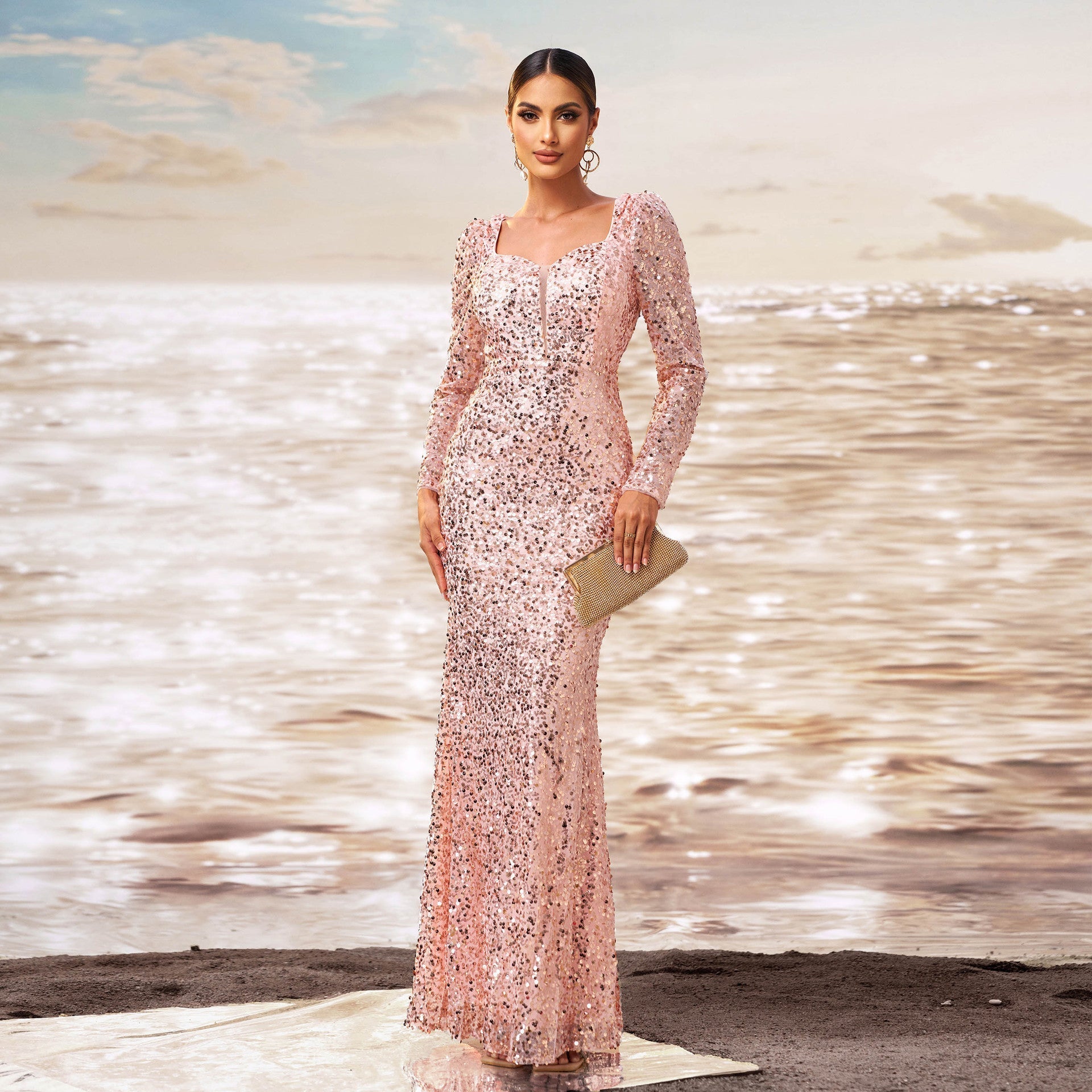 Donna in abito da sera rosa scintillante, con pochette dorata, in piedi su una spiaggia al tramonto. Eleganza, moda, abito da cerimonia.