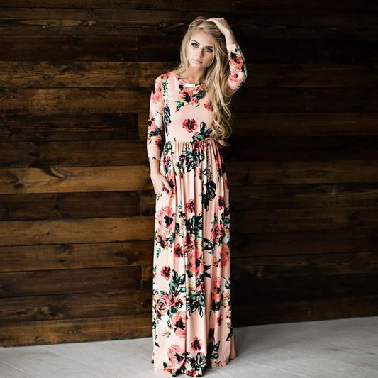 Donna con abito lungo floreale rosa, posa davanti a parete in legno scuro. Moda femminile elegante, abbigliamento primaverile, stile casual chic.