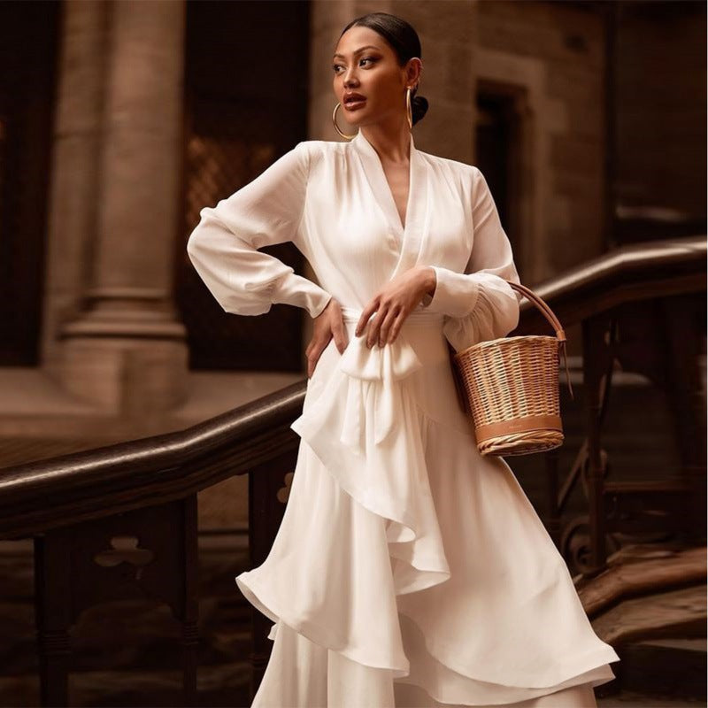 Donna elegante in abito bianco fluente con maniche lunghe, posa su scala storica. Porta una borsa di vimini. Moda sofisticata, stile classico.