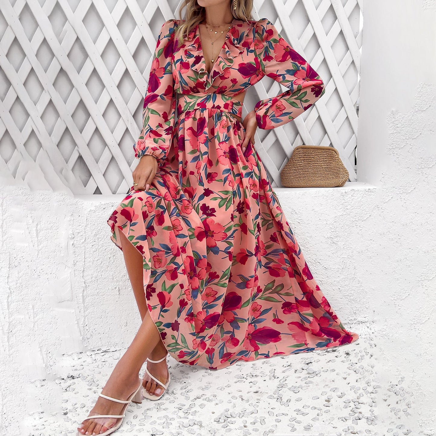Abito lungo floreale rosa e rosso, donna seduta su panca bianca, sandali bianchi, borsa paglia, sfondo grigliato bianco. Moda estiva elegante.