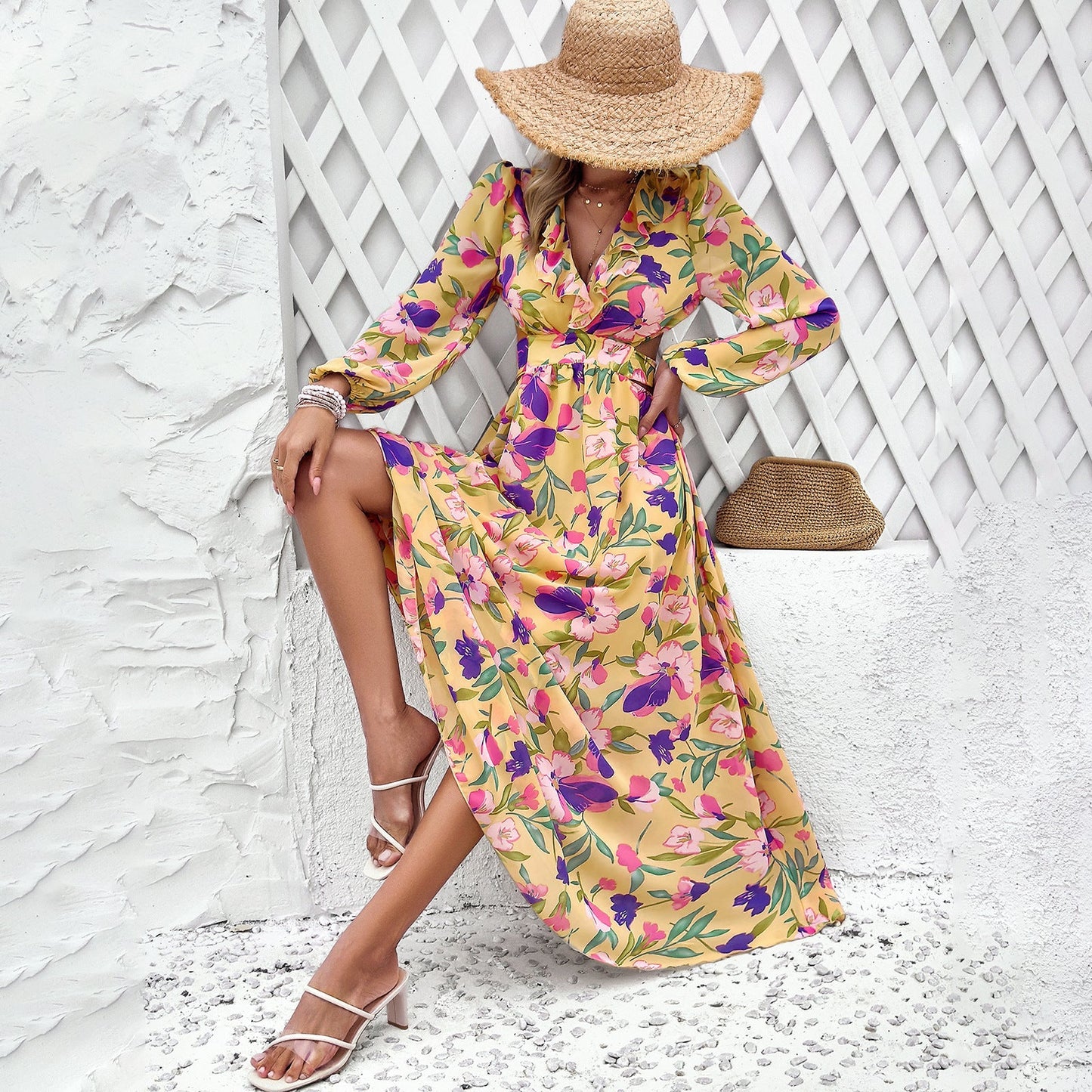 Donna con abito lungo floreale giallo, cappello di paglia, sandali bianchi, seduta su sfondo bianco. Moda estiva, stile elegante, abbigliamento femminile.