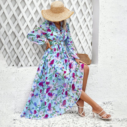 Donna con abito lungo floreale blu, cappello di paglia e sandali bianchi, seduta su sfondo bianco. Moda estiva elegante e femminile.