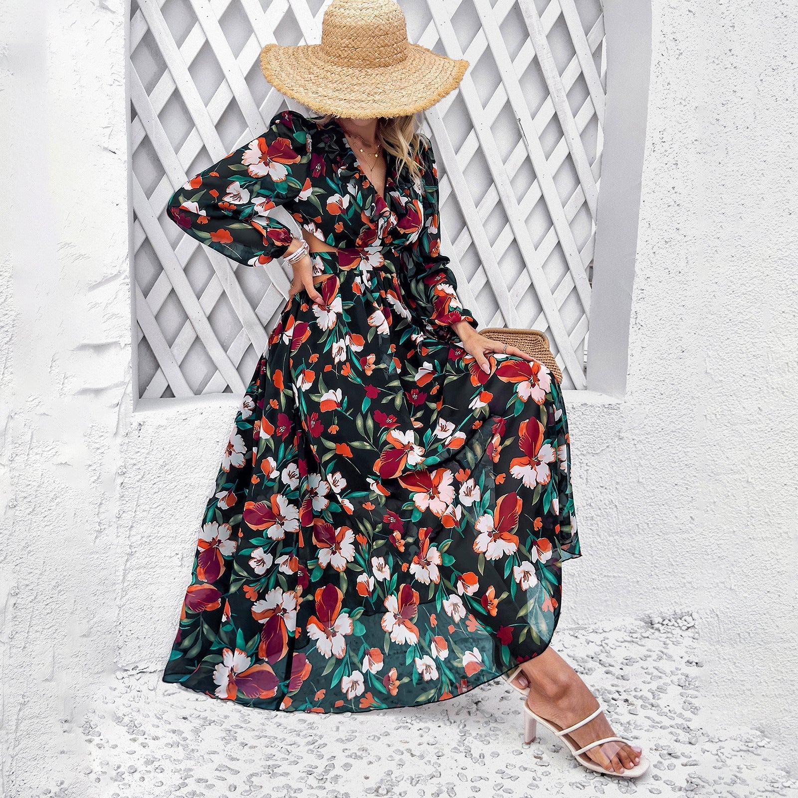 Donna con abito lungo floreale, cappello di paglia, sandali bianchi, seduta su sfondo bianco. Moda estiva, abbigliamento femminile elegante.