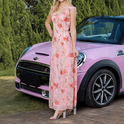 Donna in abito floreale rosa accanto a Mini Cooper rosa, su strada con alberi sullo sfondo. Auto elegante, moda estiva, stile casual chic.