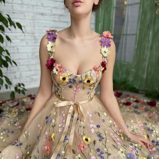 Abito floreale elegante con dettagli di fiori colorati su tessuto trasparente, indossato da una donna seduta. Sfondo con porta verde e parete bianca.