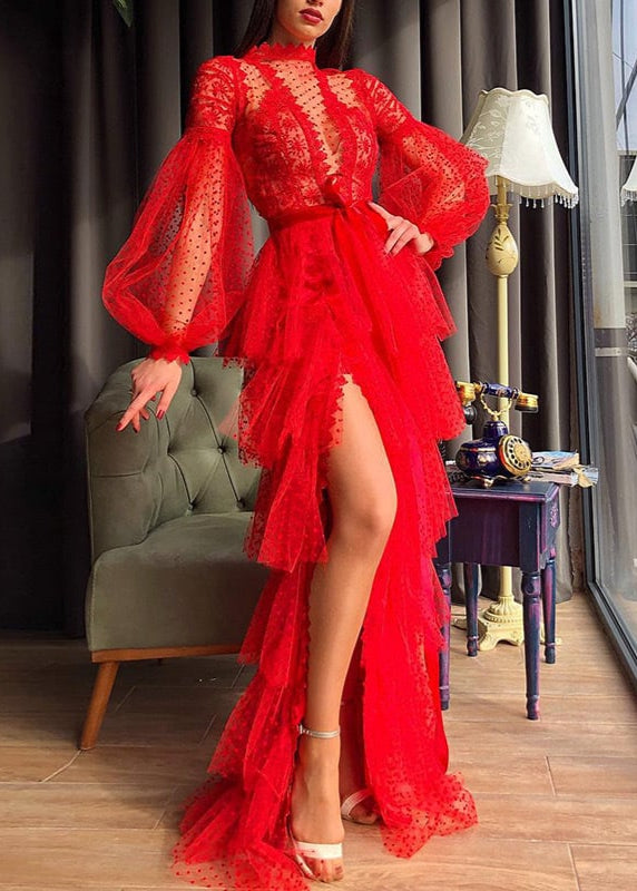 Donna in abito lungo rosso elegante con maniche a sbuffo e dettagli in pizzo, posa accanto a divano verde e lampada vintage. Moda serale glamour.