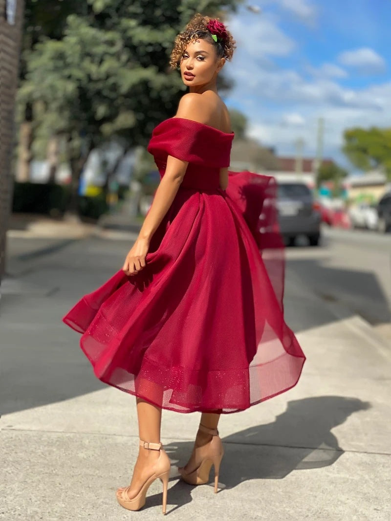 Donna in abito rosso elegante su marciapiede, sfondo urbano. Vestito off-shoulder, gonna ampia, scarpe col tacco nude. Moda femminile, stile urbano.