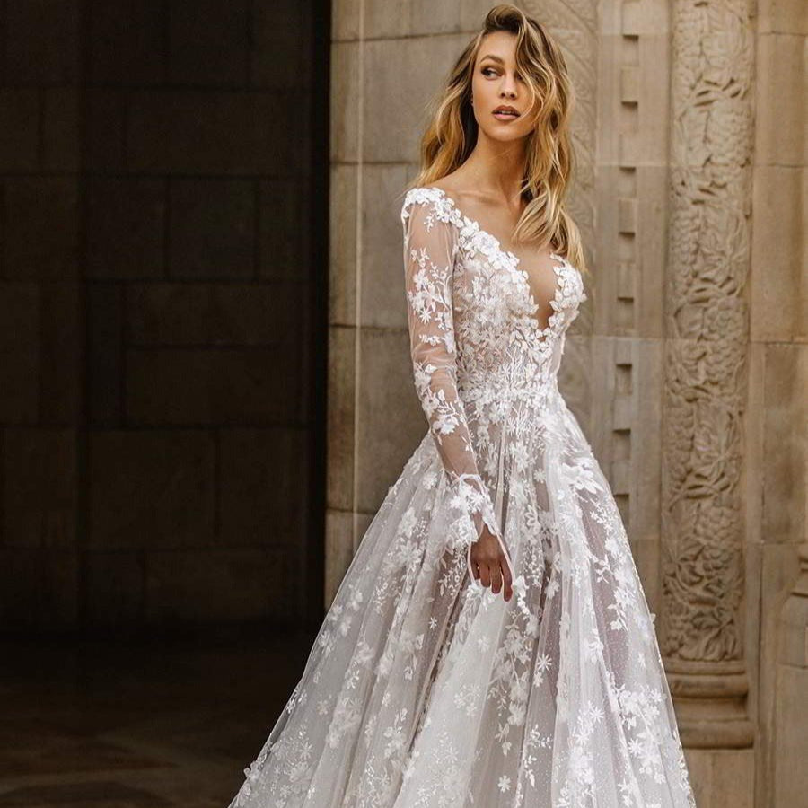 Abito da sposa elegante in pizzo floreale, maniche lunghe trasparenti, scollatura profonda, ambientato in un contesto architettonico classico.