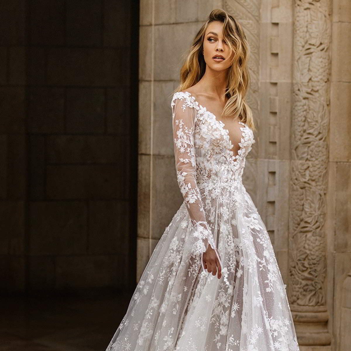 Abito da sposa elegante in pizzo bianco con dettagli floreali, maniche lunghe trasparenti e scollatura profonda, indossato da modella in ambientazione storica.