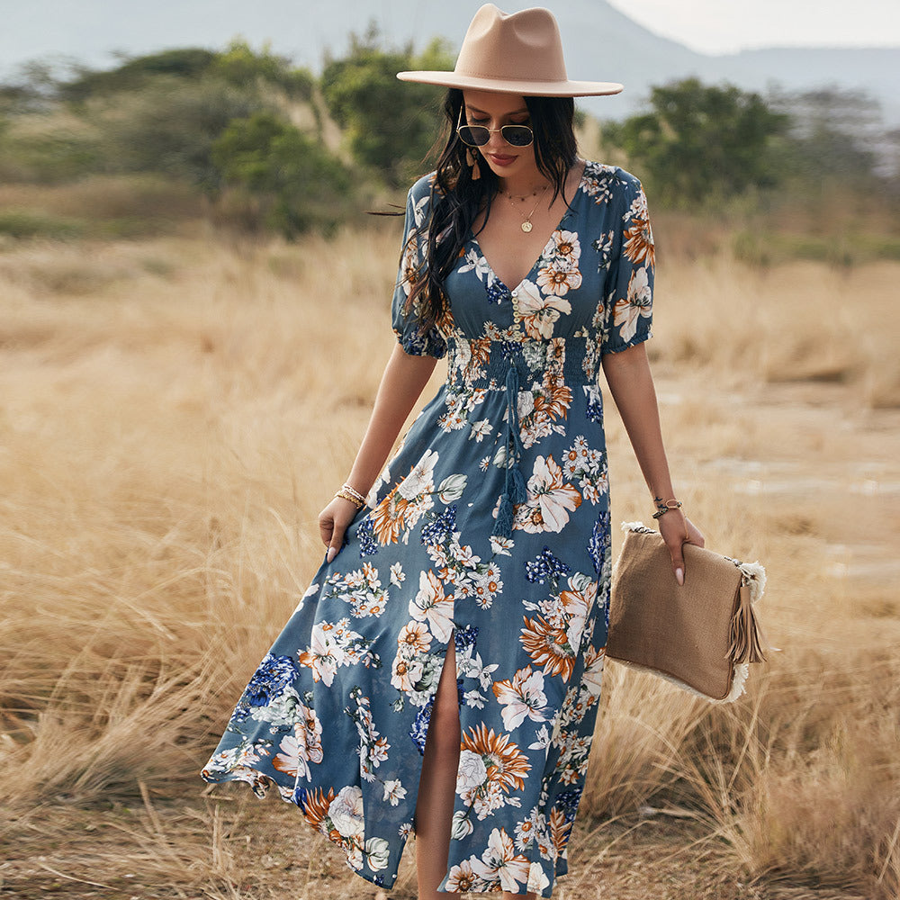 Donna con abito floreale blu, cappello beige e occhiali da sole, cammina in un campo. Moda estiva, stile boho-chic, abbigliamento casual elegante.