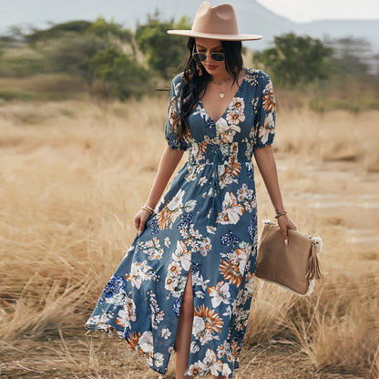 Donna con abito floreale blu, cappello beige e occhiali da sole, cammina in un campo. Moda estiva, stile boho-chic, abbigliamento casual elegante.