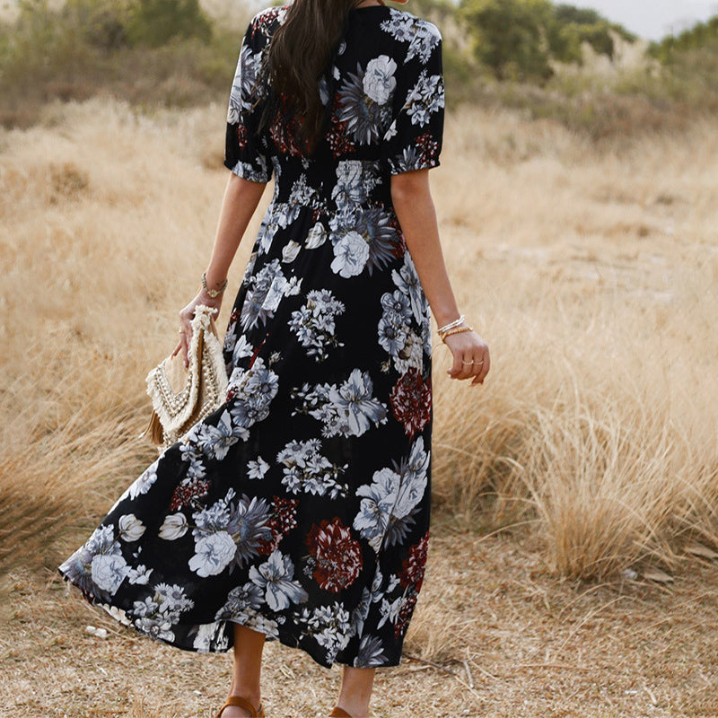 Donna in abito floreale lungo nero cammina in un campo erboso, tenendo una borsa intrecciata. Moda estiva, stile boho-chic, natura.