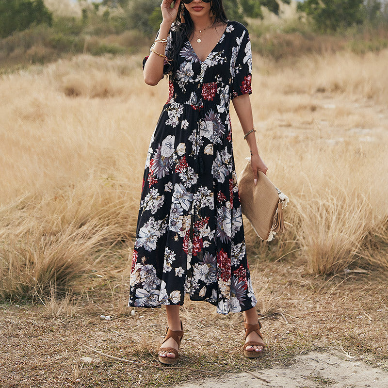 Donna con abito lungo floreale nero, sandali marroni e borsa beige in un campo erboso. Moda estiva, abbigliamento femminile elegante.