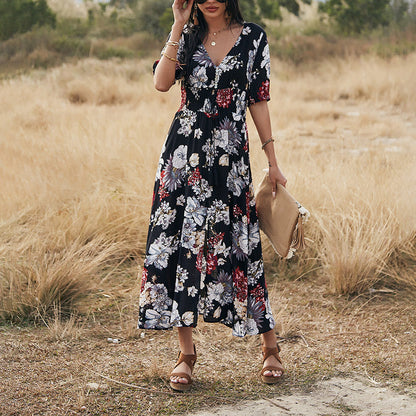 Donna con abito lungo floreale nero, sandali marroni e borsa beige in un campo erboso. Moda estiva, abbigliamento femminile elegante.