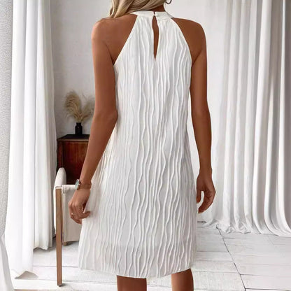 Donna in abito bianco plissettato, senza maniche, in una stanza luminosa con tende bianche e sedia moderna. Eleganza estiva, moda femminile.
