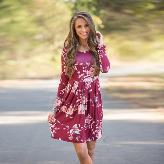 Donna sorridente con abito floreale rosso bordeaux, maniche lunghe, in un ambiente naturale. Moda autunnale, abbigliamento femminile elegante.