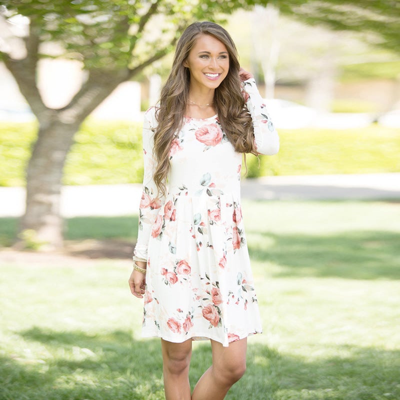 Donna sorridente con abito floreale bianco e rosa, passeggia in un parco verdeggiante. Moda primaverile, abbigliamento casual elegante.