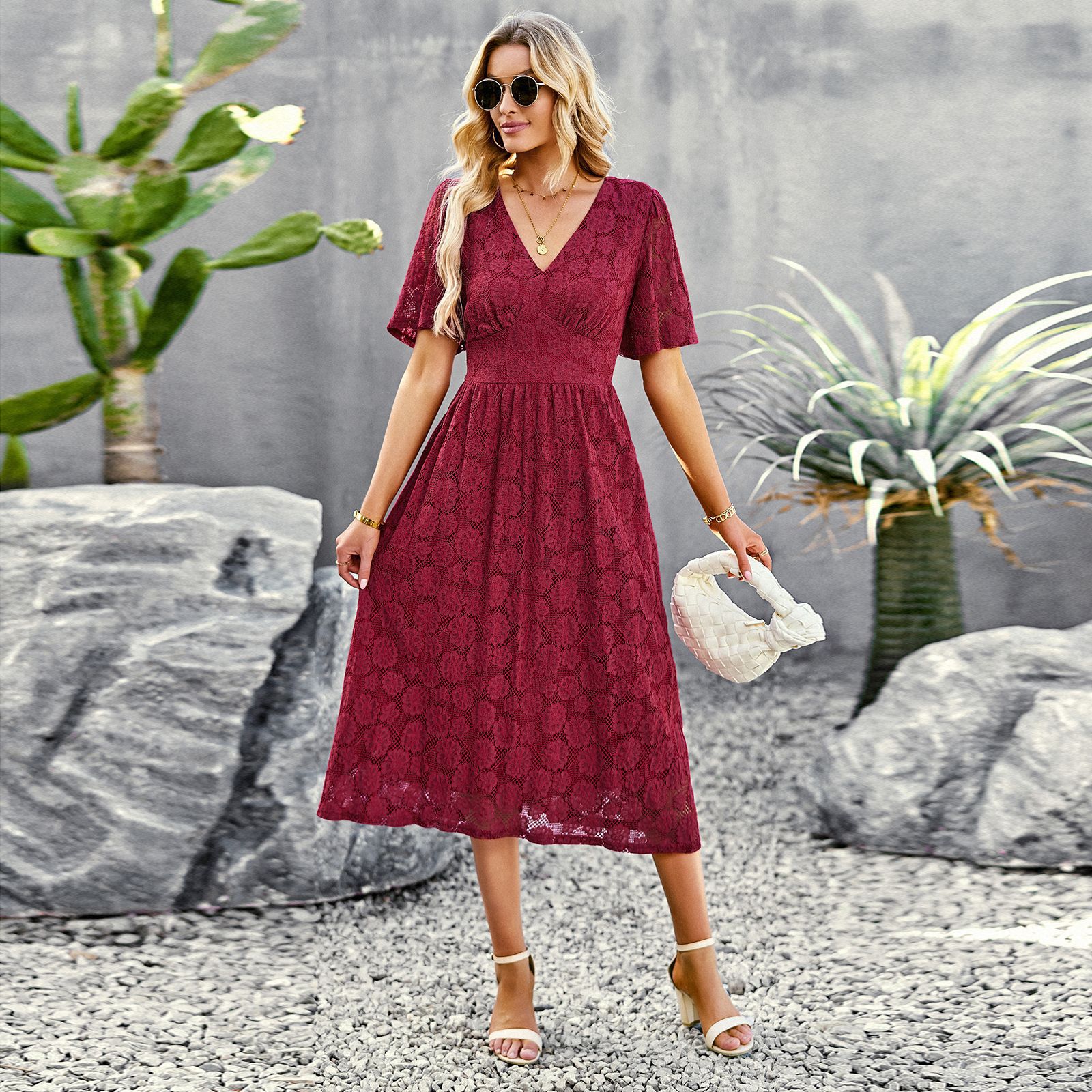 Donna con abito rosso in pizzo, occhiali da sole, sandali bianchi, borsa bianca, in giardino con cactus e piante tropicali. Moda estiva elegante.