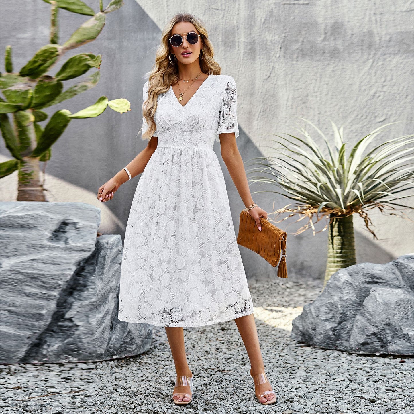 Donna in abito bianco di pizzo, occhiali da sole, borsa marrone, sandali, posa in giardino con piante succulente e pietre decorative. Moda estiva elegante.